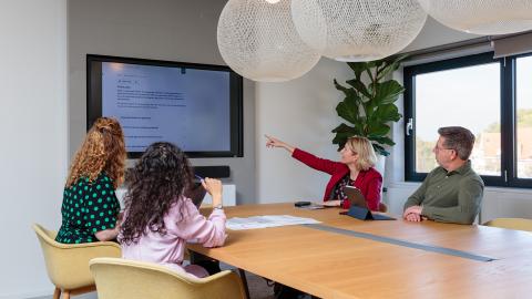 In de vergaderkamer zitten collega's in overleg en op het scherm zie je een aantal collega's die digitaal deelnemen aan de vergadering