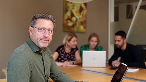 Een collega kijkt richting de camera terwijl de rest van het team op de achtergrond verder aan het vergaderen is