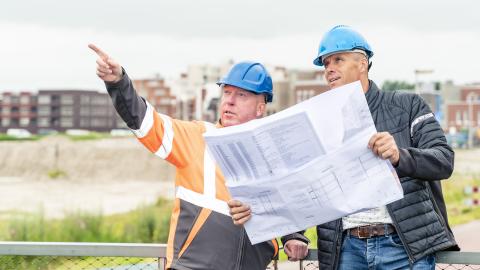Twee collega's staan op de Melkwegbrug en kijken naar een groot vel met plannen. Op de achtergrond is de Weidevenne zichtbaar